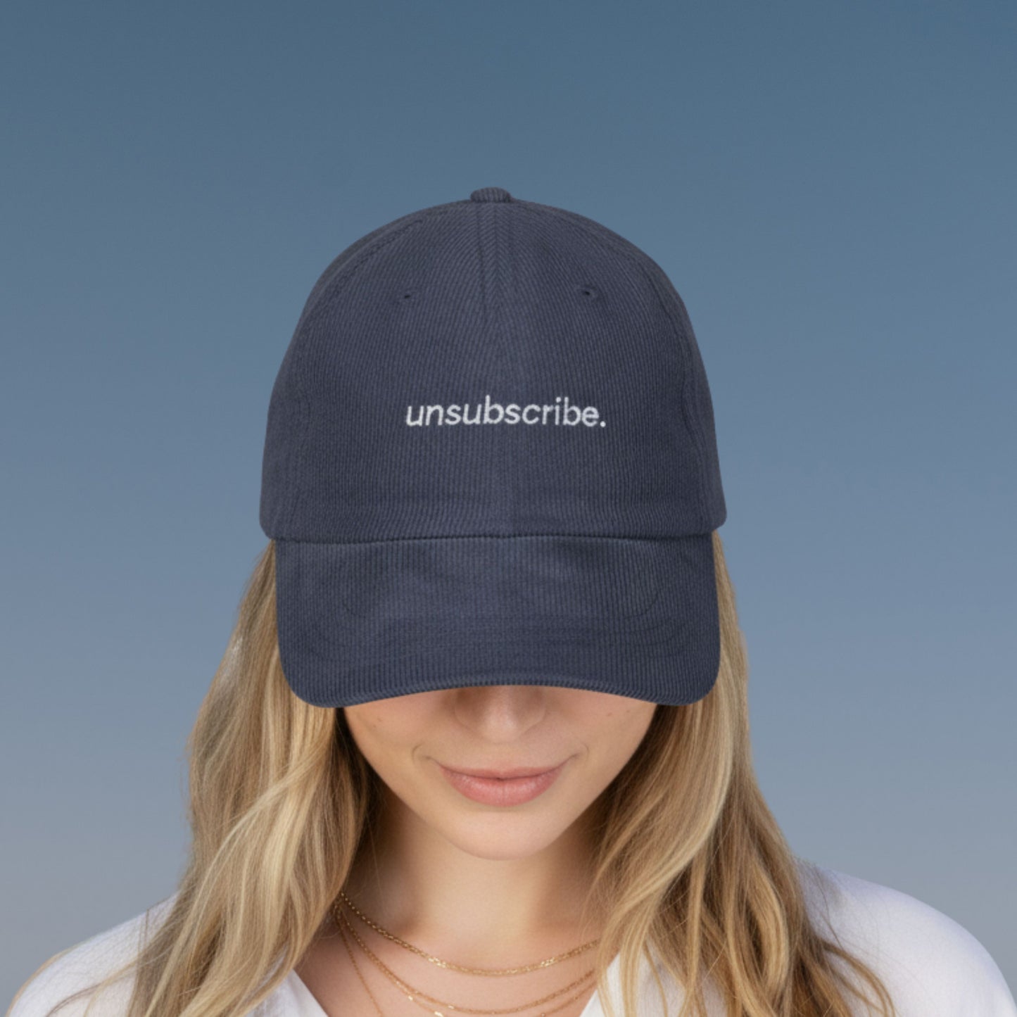Person wearing a navy blue corduroy cap with 'unsubscribe.' text embroidered on a gradient blue background