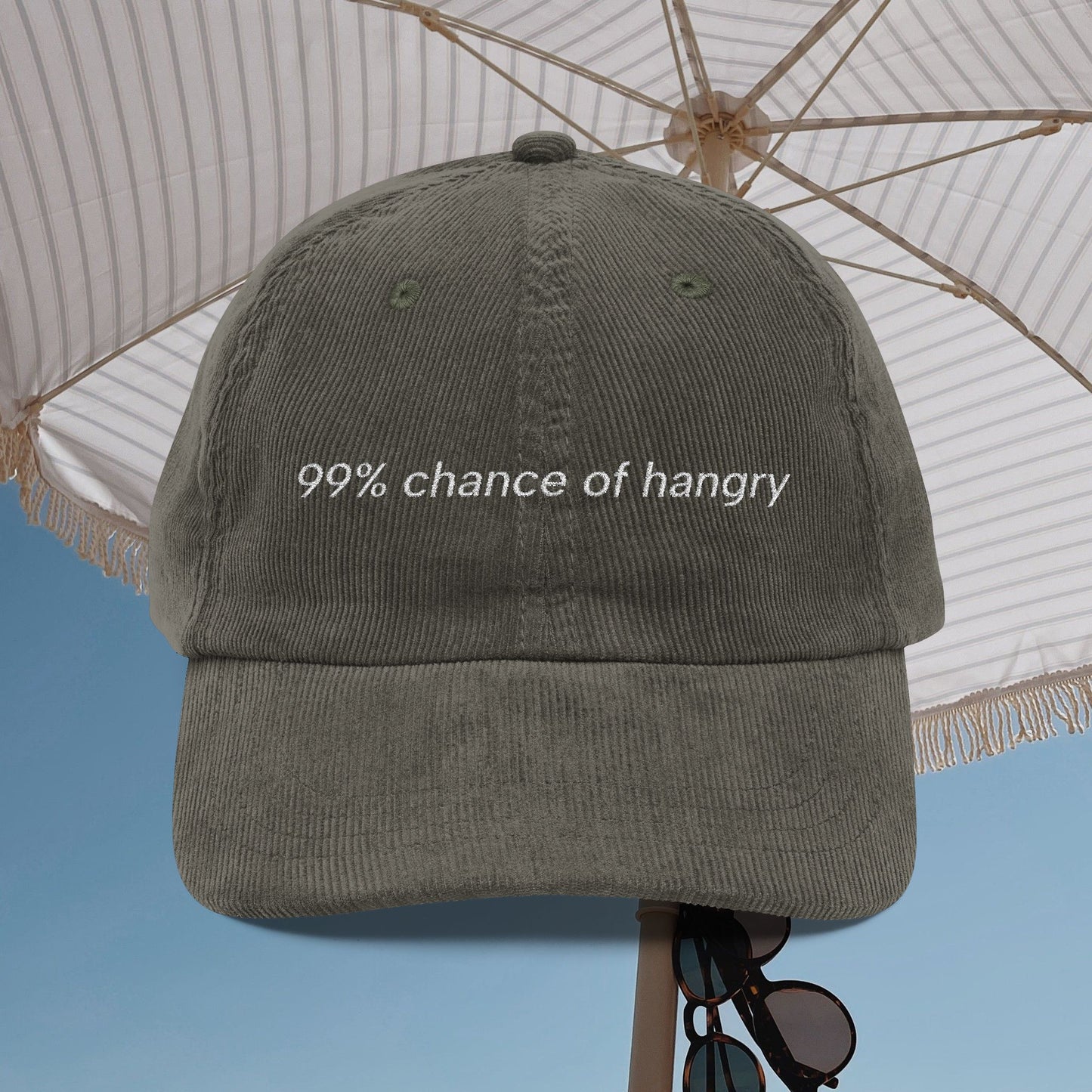 Gray corduroy cap with '99% chance of hangry' embroidered text under a white umbrella with blue sky background
